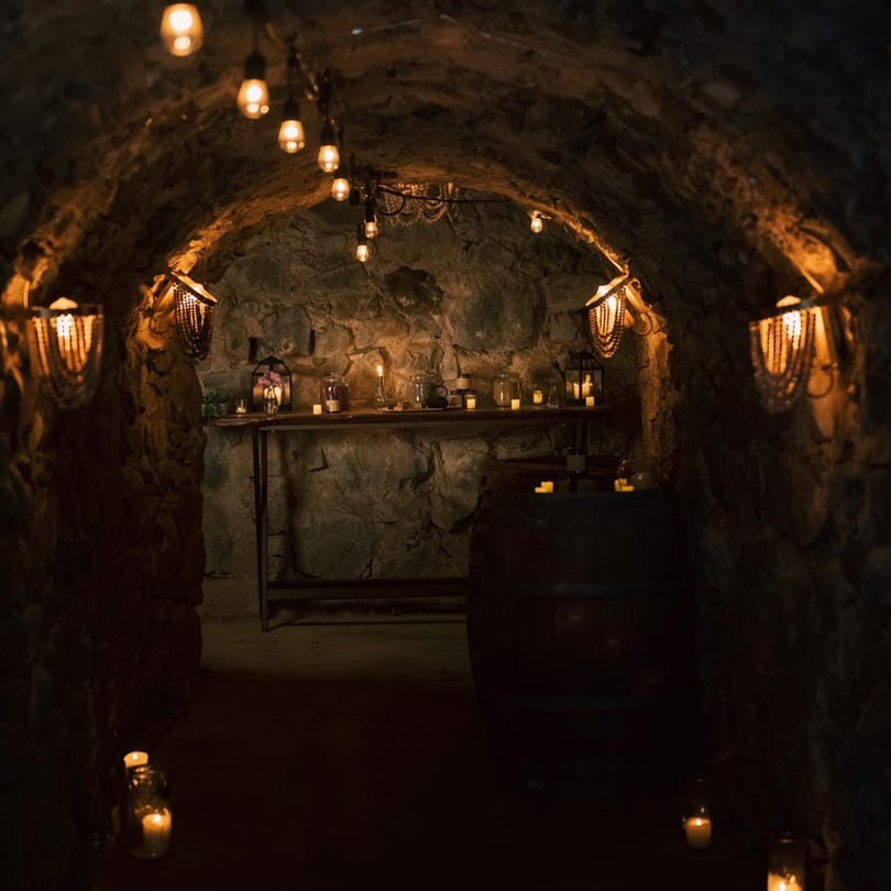 A dimly lit stone cellar or tunnel with string lights, wall sconces, and candles creating a warm ambiance.