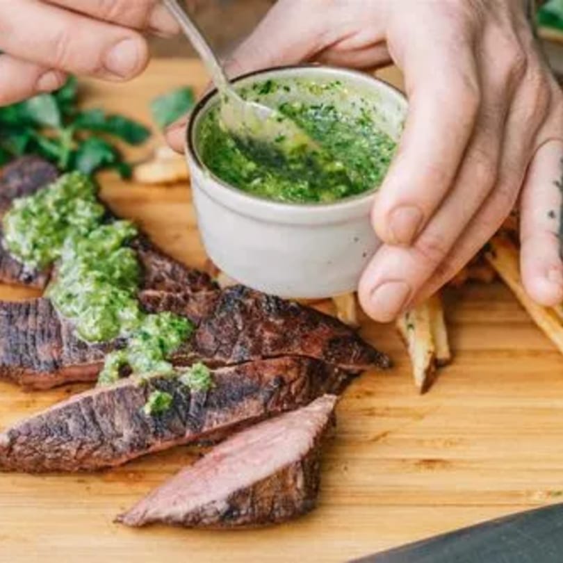 Hands spooning vibrant green chimichurri sauce over sliced New American steak on a wooden board.