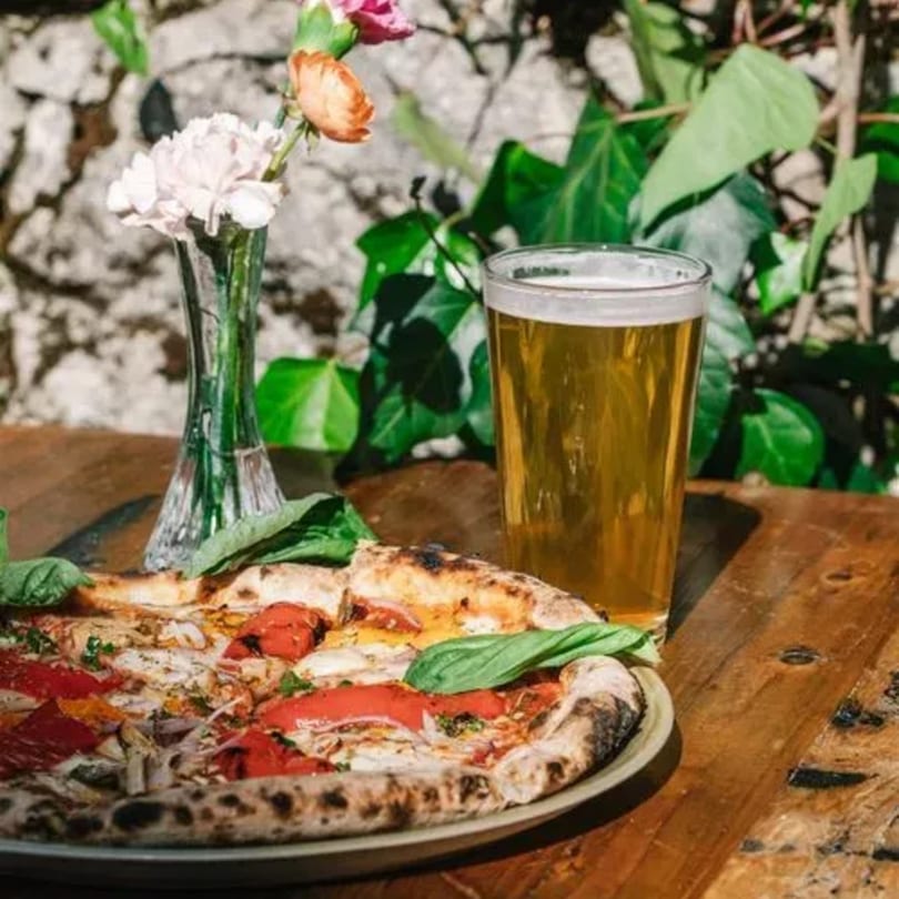 Stone House pizza with red peppers and basil, a beer, and flowers on a wooden table.