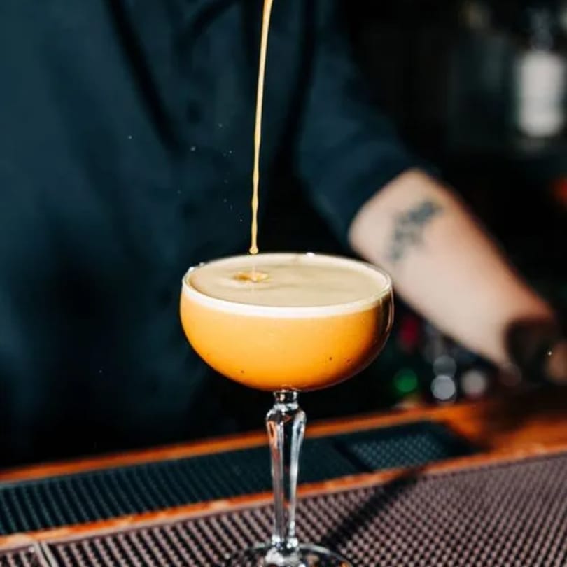 A bartender pours a golden cocktail into a coupe glass on a dark bar surface at Stone House.