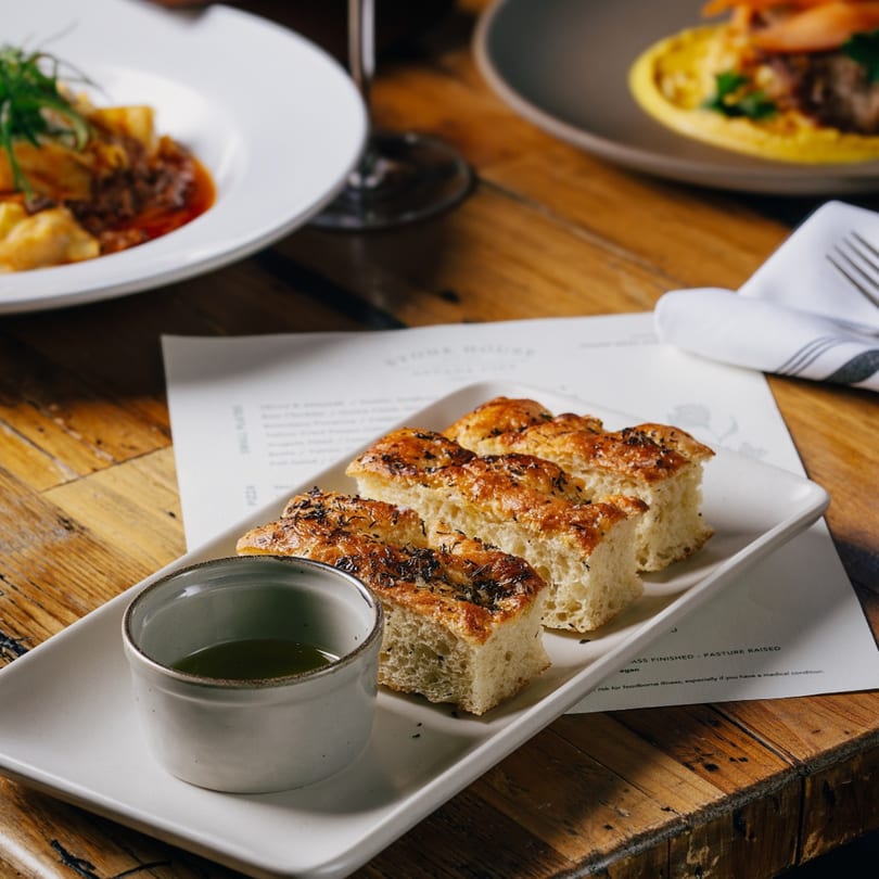 Focaccia bread with herbs and olive oil dip, with other dishes blurred in the background.