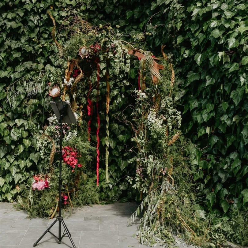 A lush green floral arch with red and white flowers, ribbons, and a black tripod stand.