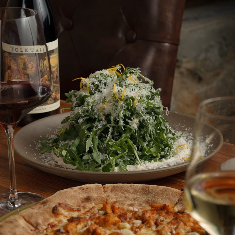 Arugula salad with parmesan and lemon zest, pizza, and wine glasses on a wooden table.