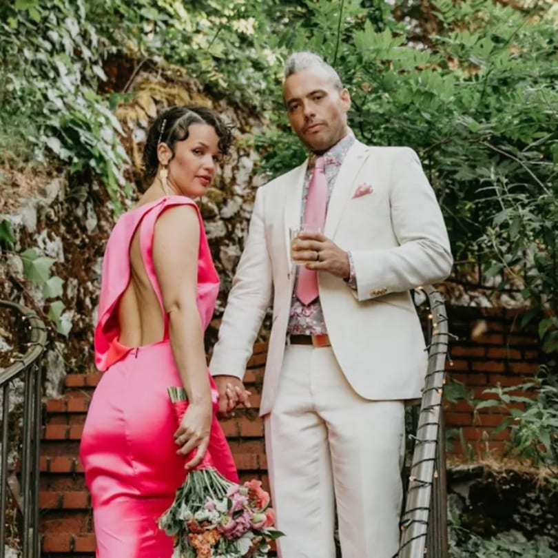 A couple in formal attire, the woman in a pink dress and the man in a white suit, on outdoor stairs.