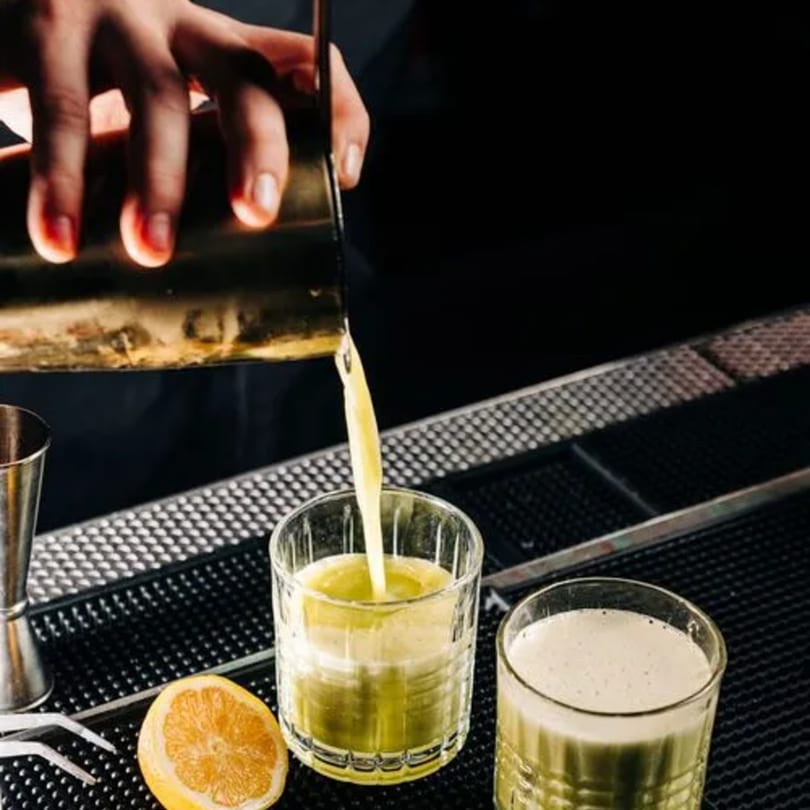 Bartender pouring a green cocktail from a shaker into a glass on a dark bar counter.