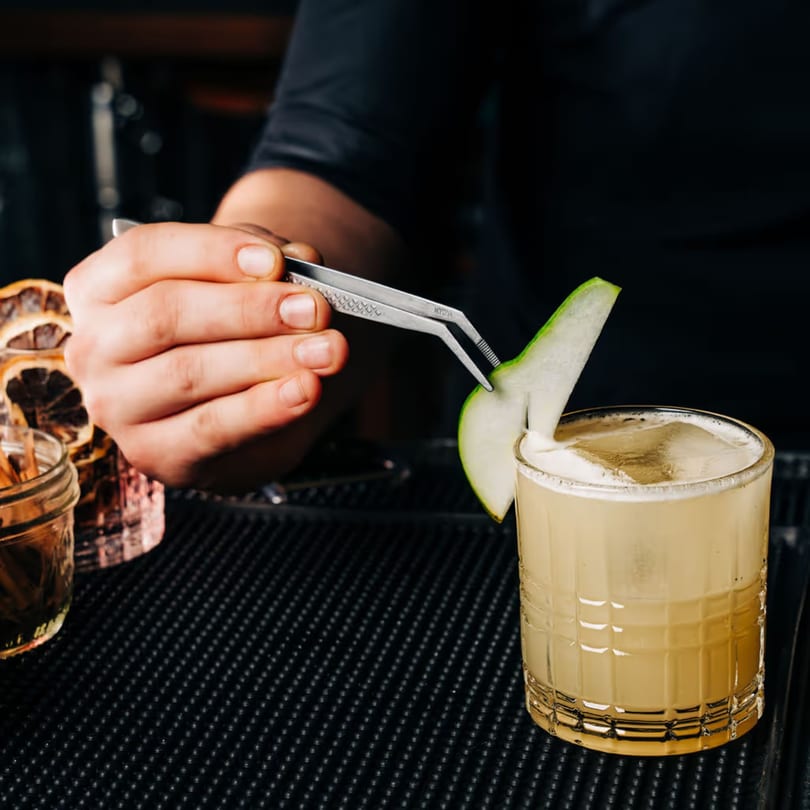 A bartender garnishes a Stone House cocktail with a green apple slice using tweezers.