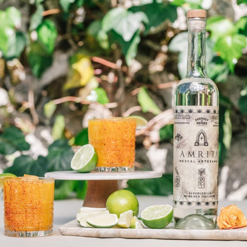 Two mezcal cocktails with chili-lime rims, limes, and a bottle of Amrita Mezcal Artesanal.