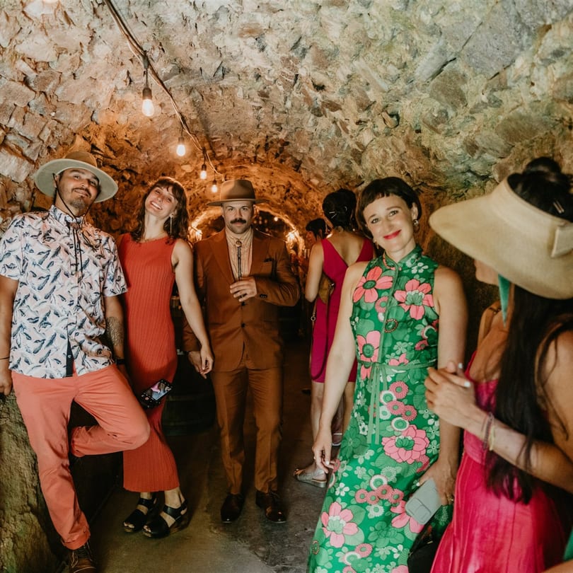 Guests in stylish attire pose in a dimly lit, stone-walled tunnel with string lights.