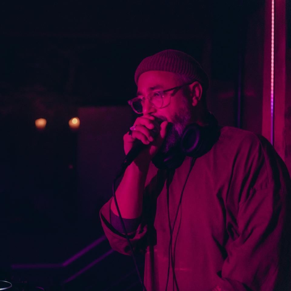 A man in glasses and a beanie sings into a microphone under pink lighting at Stone House.