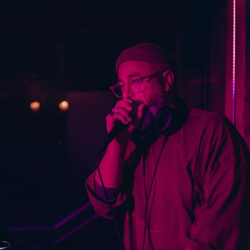 A man in glasses and a beanie sings into a microphone under pink lighting at Stone House.
