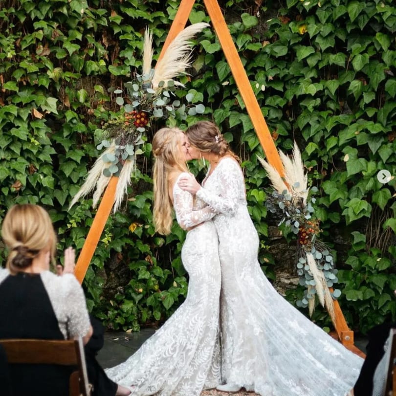 Two brides in white lace gowns kiss under a wooden arch adorned with pampas grass and eucalyptus.