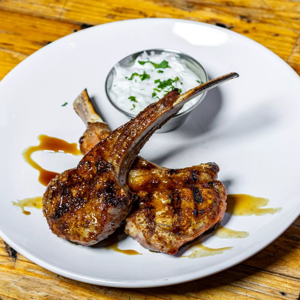 Two grilled lamb chops with a drizzle of sauce, served with a side of tzatziki on a white plate.