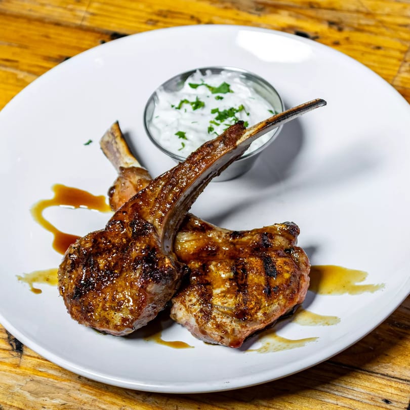 Two grilled lamb chops with a drizzle of sauce, served with a side of tzatziki on a white plate.