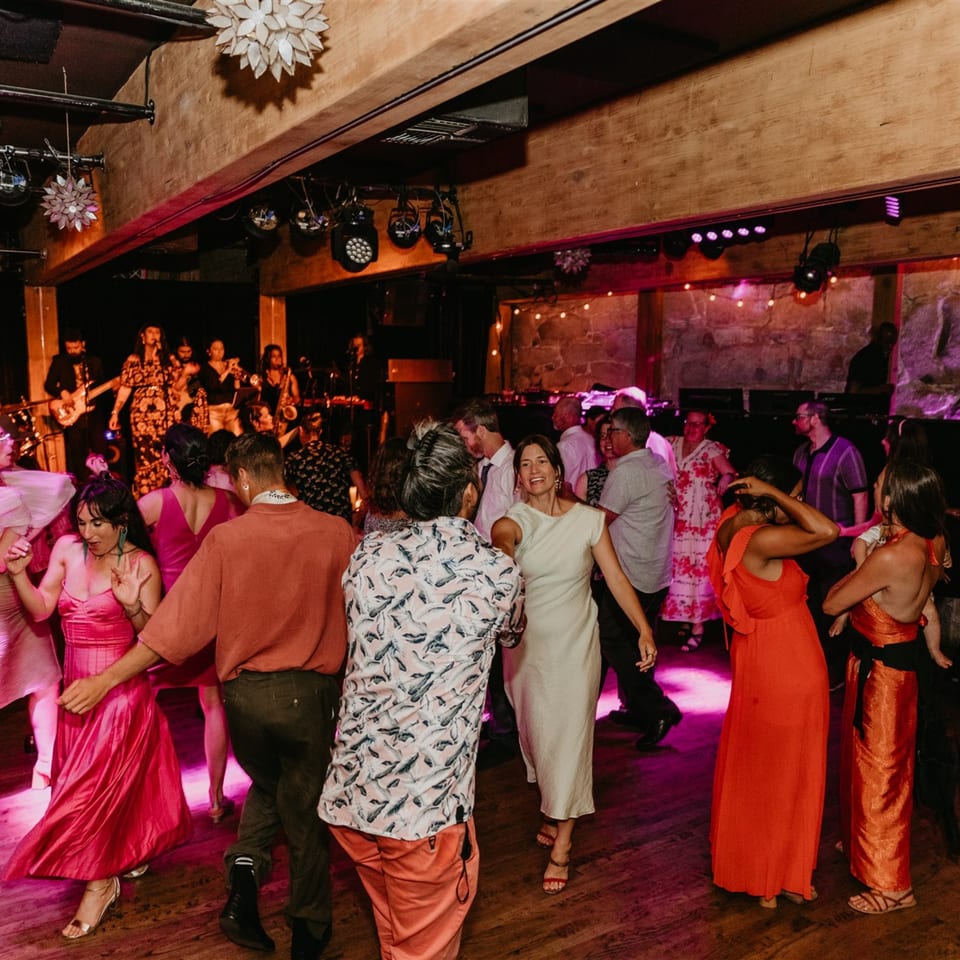 Guests dancing on a wooden floor under pink lights, with a band performing in the background.