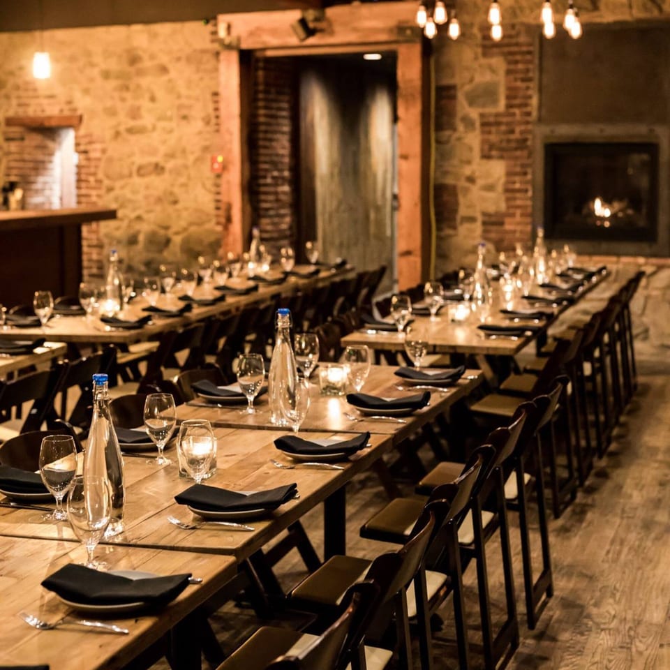 Long wooden tables set for a banquet with black chairs, stone walls, and a fireplace at Stone House.