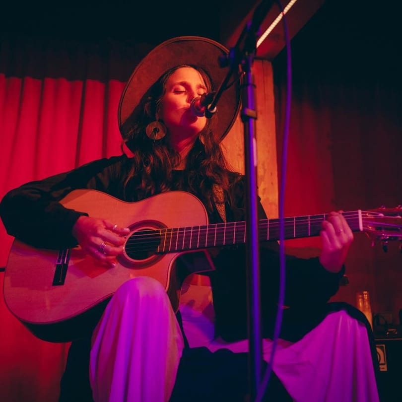 A musician with a hat sings into a microphone while playing an acoustic guitar on a red-lit stage.