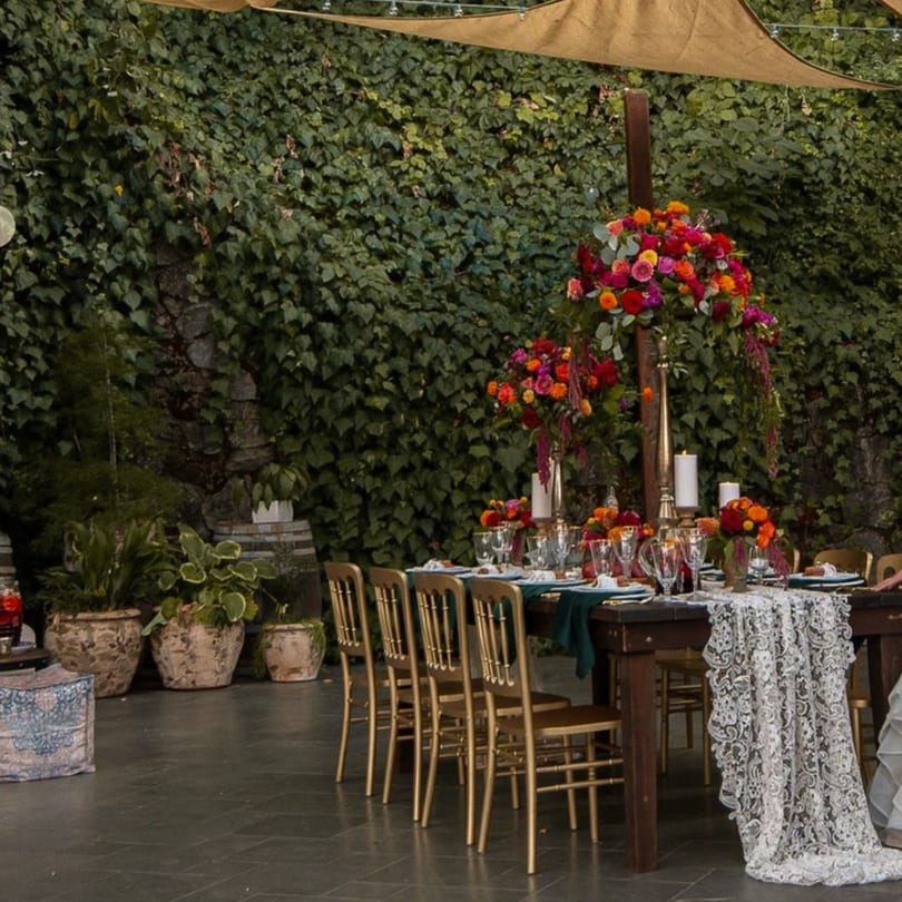An outdoor dining table set with vibrant floral centerpieces, gold chairs, and lace runner against an ivy wall.