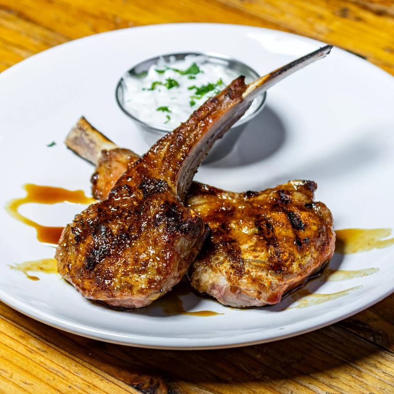 Two grilled lamb chops with a side of white sauce and green garnish on a white plate.