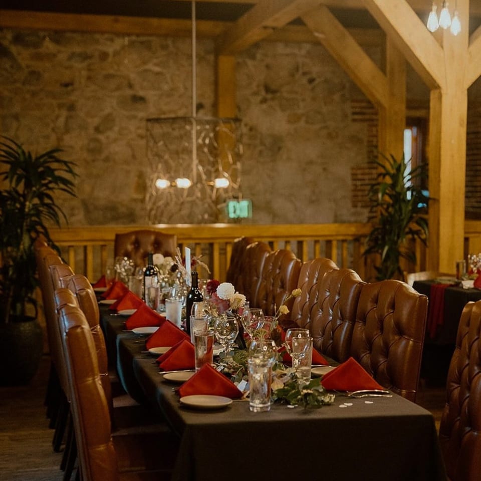 Long dining table set with red napkins, wine bottles, and floral centerpieces in a rustic room.