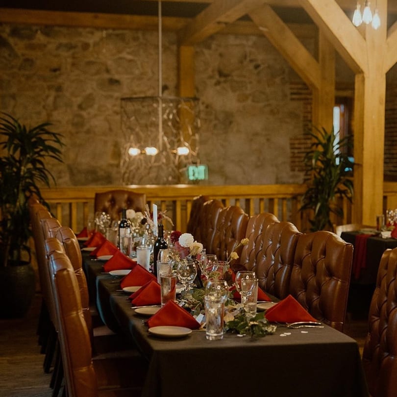 Long dining table set with red napkins, wine bottles, and floral centerpieces in a rustic room.