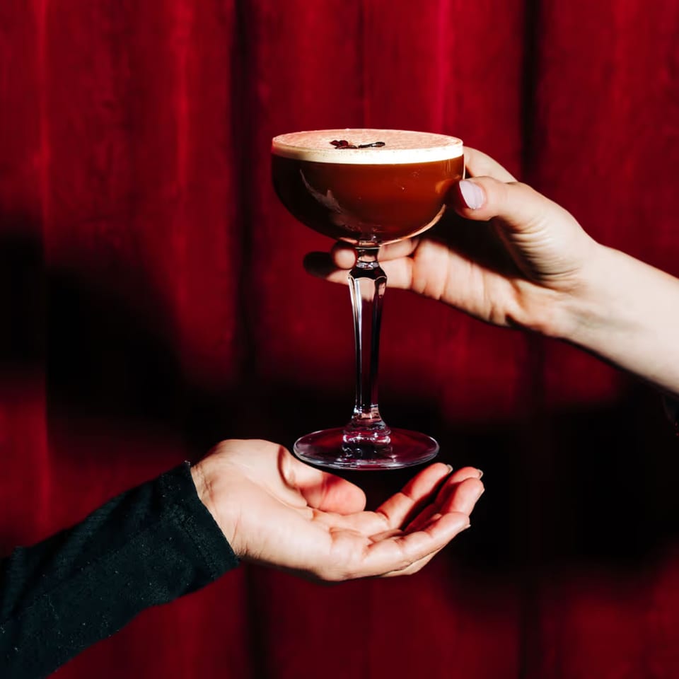 A hand holding a dark cocktail with foam and coffee beans, passed to another hand against a red background.