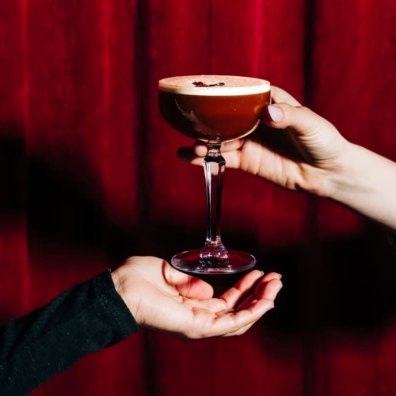 A hand holding a dark cocktail with foam and coffee beans, passed to another hand against a red background.