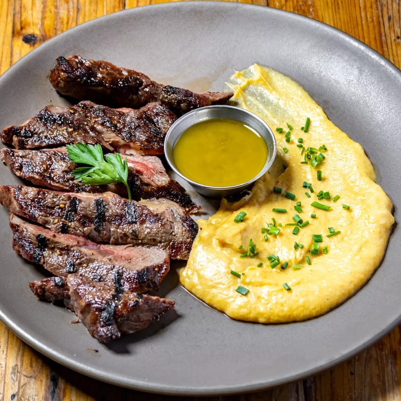 Grilled steak slices with a side of yellow mashed potatoes, green sauce, and parsley on a gray plate.