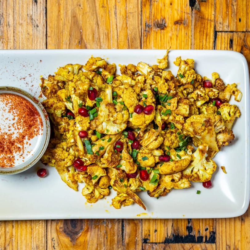 Roasted cauliflower with pomegranate seeds, green onions, and a side of white sauce with paprika.