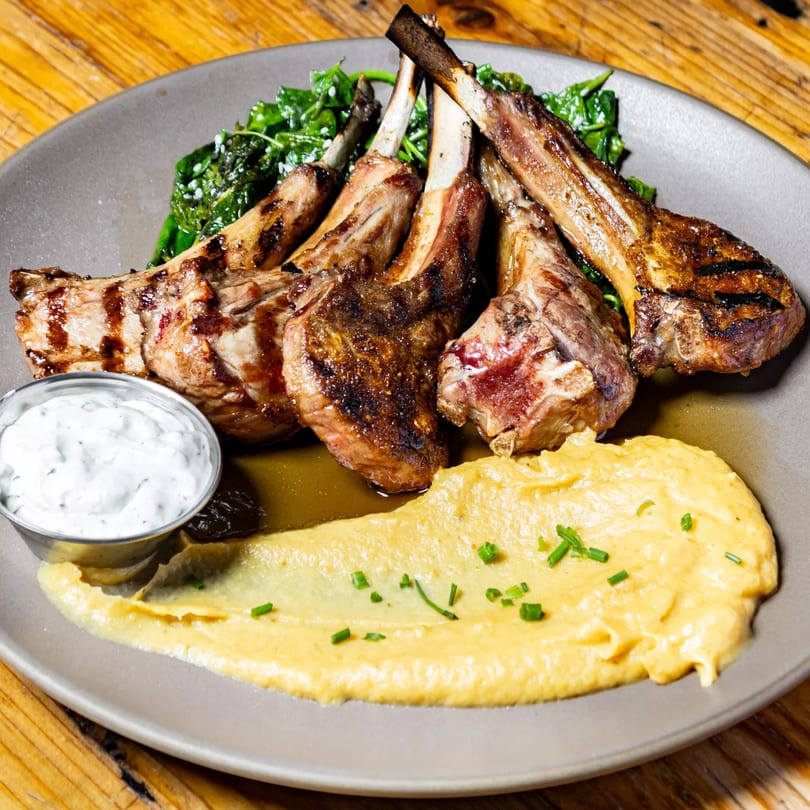 Grilled lamb chops with a side of creamy polenta, sautéed greens, and a white dipping sauce.