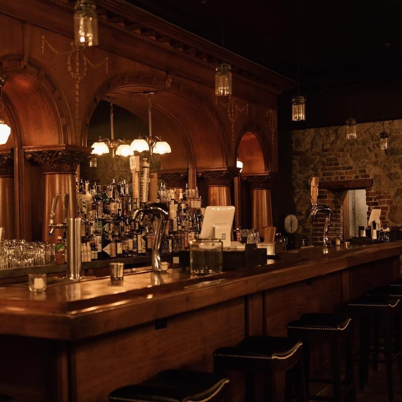 A dimly lit, elegant bar at Stone House with a long wooden counter, stools, and many liquor bottles.