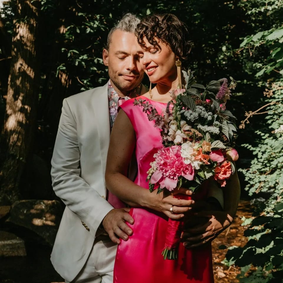 A couple embracing outdoors, the woman in a pink dress holding a large floral bouquet.