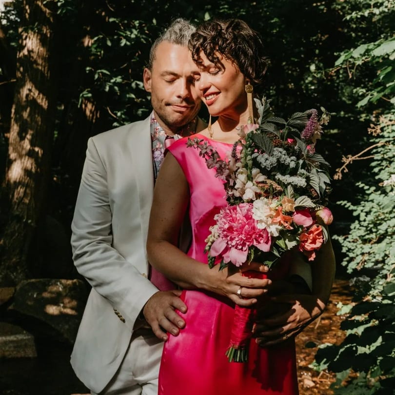 A couple embracing outdoors, the woman in a pink dress holding a large floral bouquet.