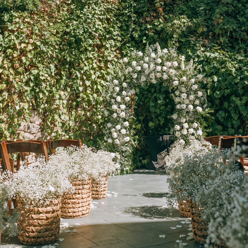 Outdoor wedding ceremony setup with white floral arch, wooden chairs, and baby's breath in woven baskets.
