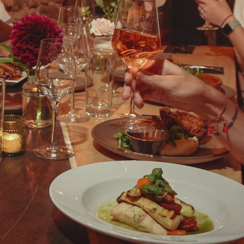 A person holding a glass of rosé wine at a table with New American dishes and floral decor.