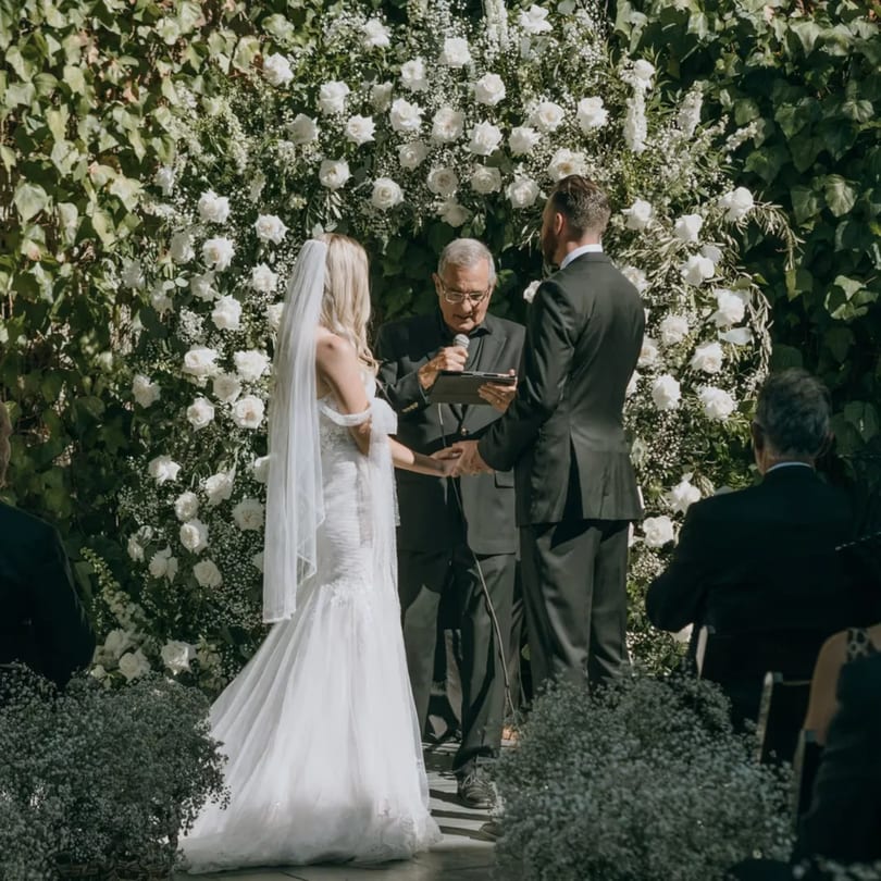 A wedding ceremony with a couple holding hands before an officiant, backed by a white floral arch.