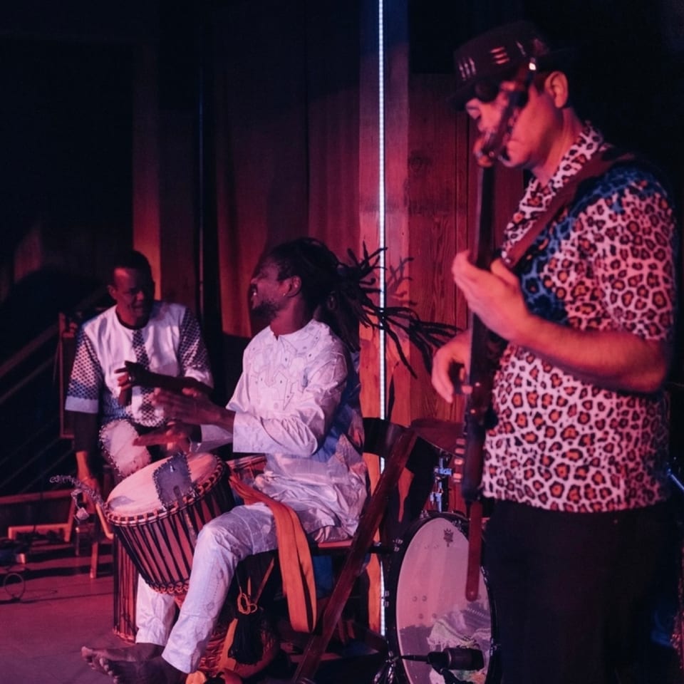 Three musicians performing with drums and a guitar on a dimly lit stage at Stone House.