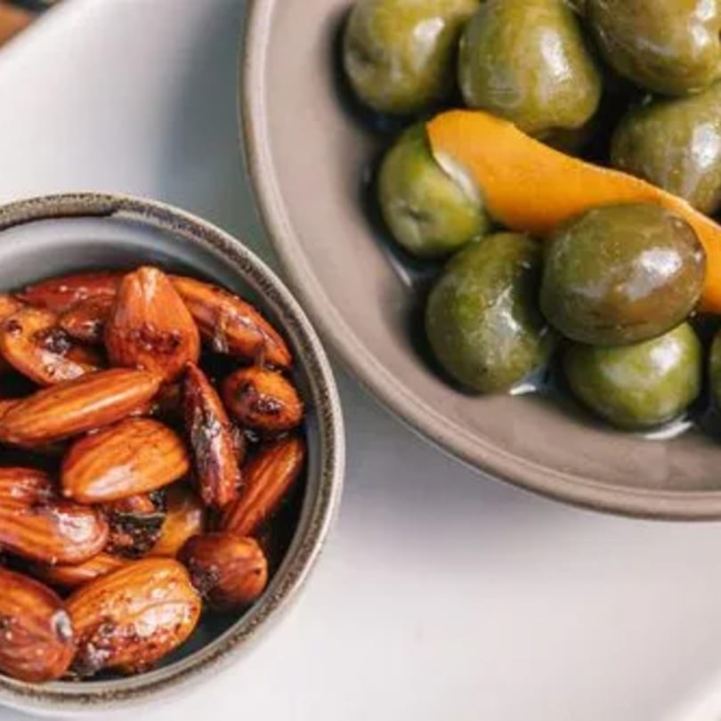 Two bowls of appetizers: roasted almonds in the foreground and green olives with orange peel.