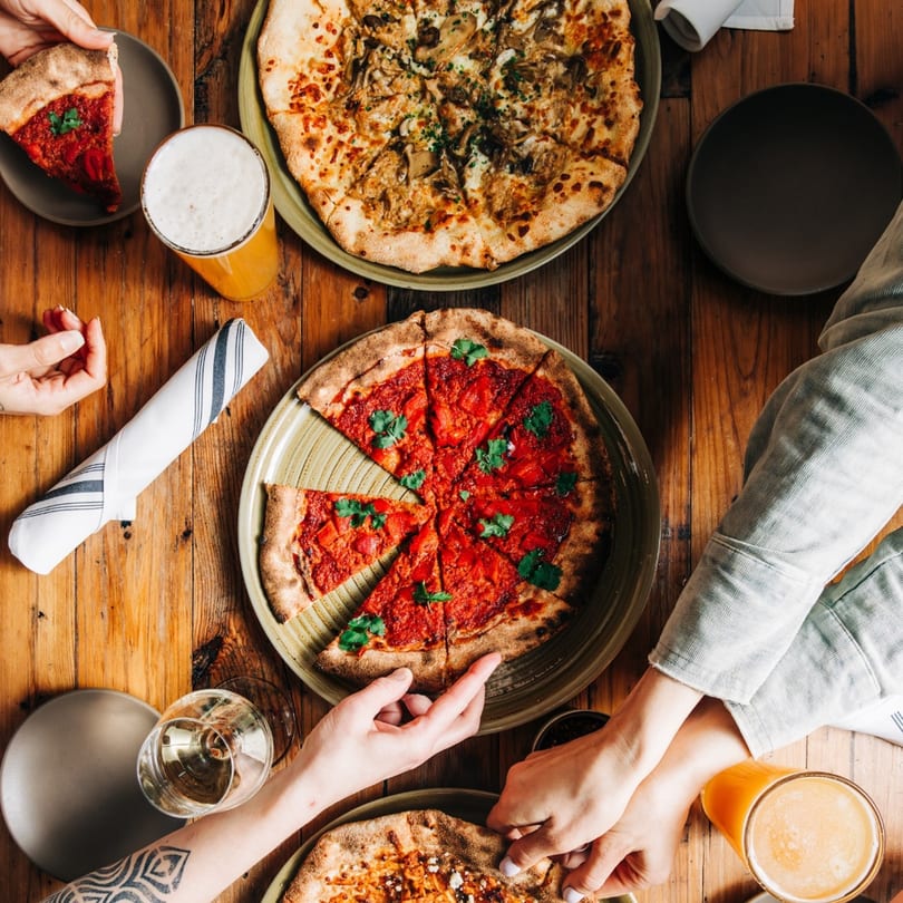Overhead view of hands reaching for slices of New American pizzas and drinks on a wooden table.