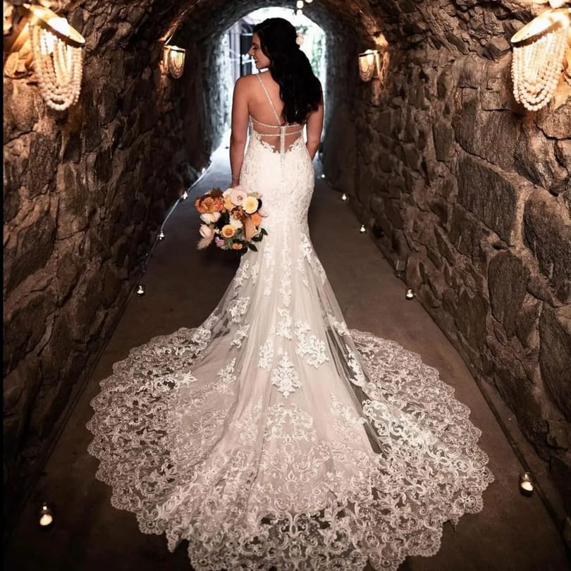 A bride in a white lace gown with a long train walks through a stone archway.