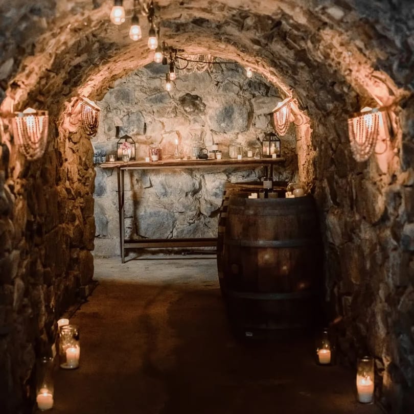 A dimly lit stone archway with string lights, wall sconces, candles, and a wooden barrel.