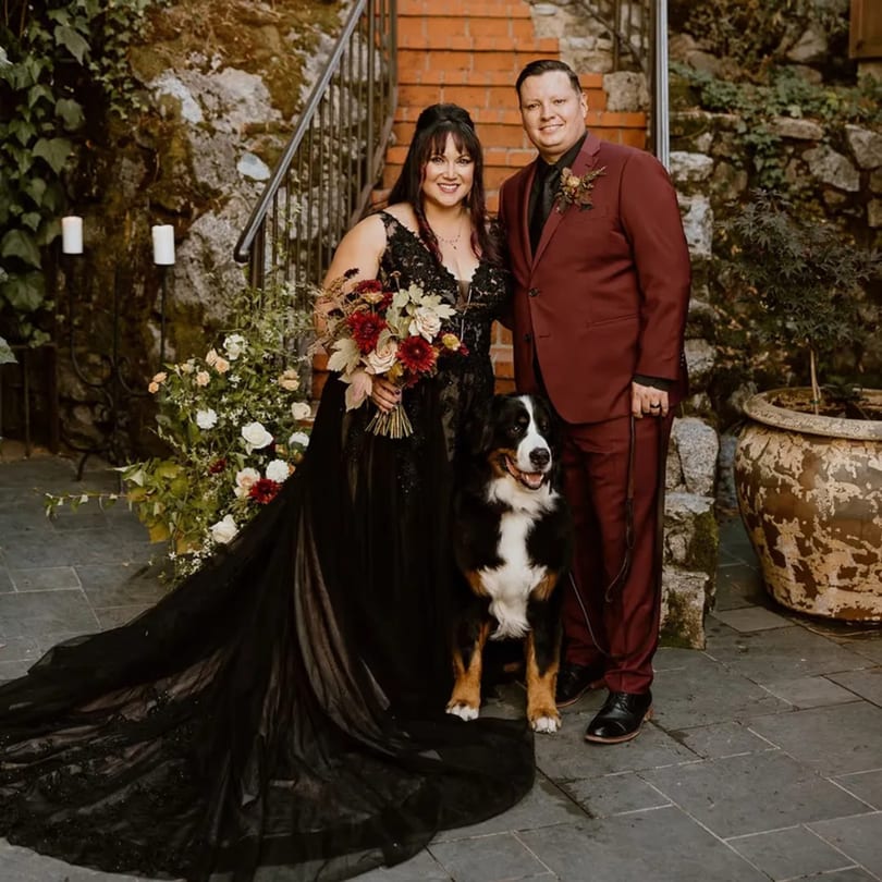 A couple in wedding attire with their Bernese Mountain Dog, standing on a patio with stone walls.