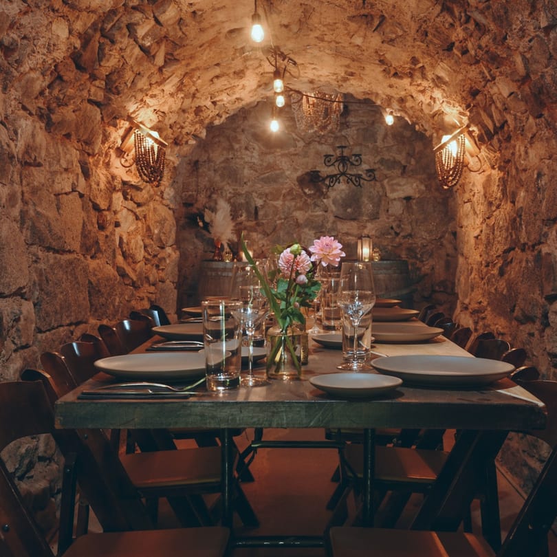 A long dining table set with plates, glasses, and flowers in a cozy, stone-walled room.