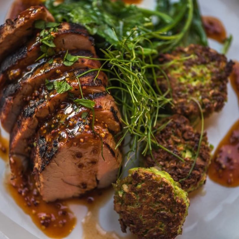 Sliced grilled pork tenderloin with mustard sauce, green vegetables, and crispy fritters on a white plate.