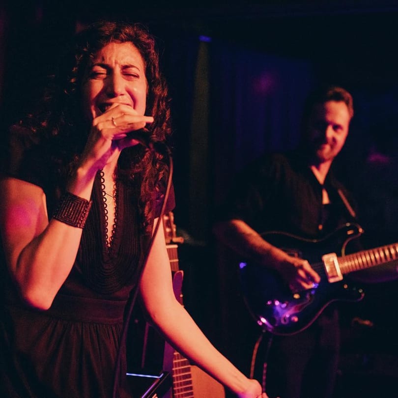 A female singer with curly hair and a male guitarist performing live on a dimly lit stage.