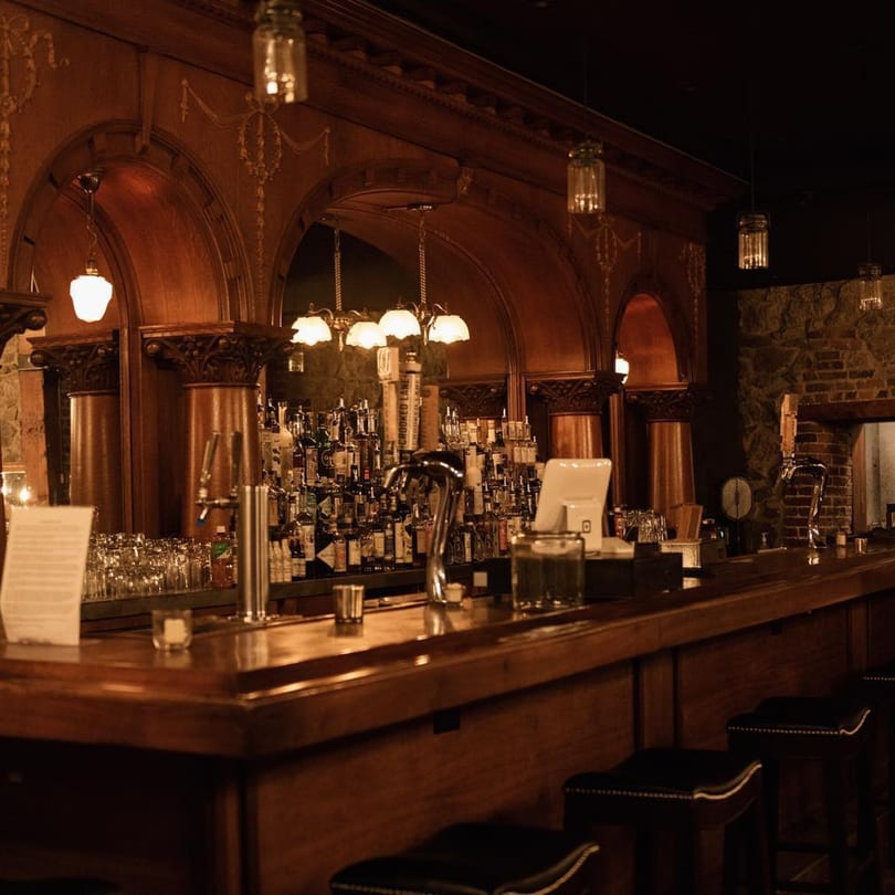 A dimly lit, elegant wooden bar with bottles, glasses, and bar stools at Stone House restaurant.