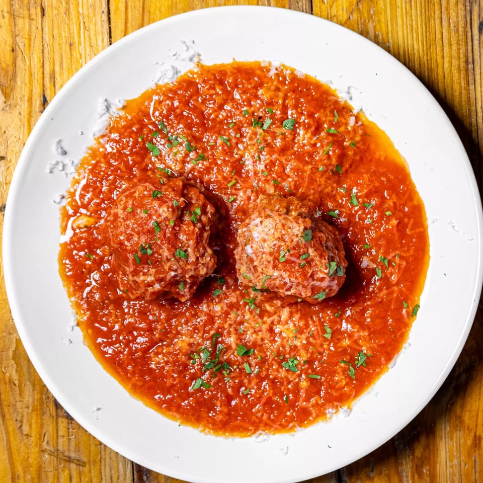 Two meatballs in rich red sauce, topped with grated cheese and fresh parsley, on a white plate.