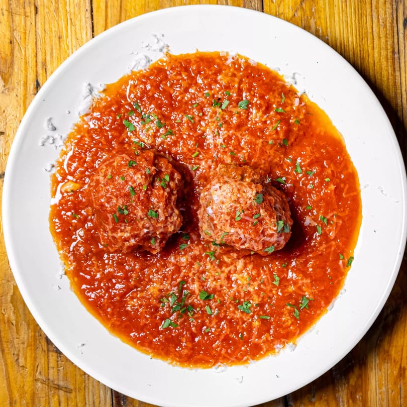 Two meatballs in rich red sauce, topped with grated cheese and fresh parsley, on a white plate.