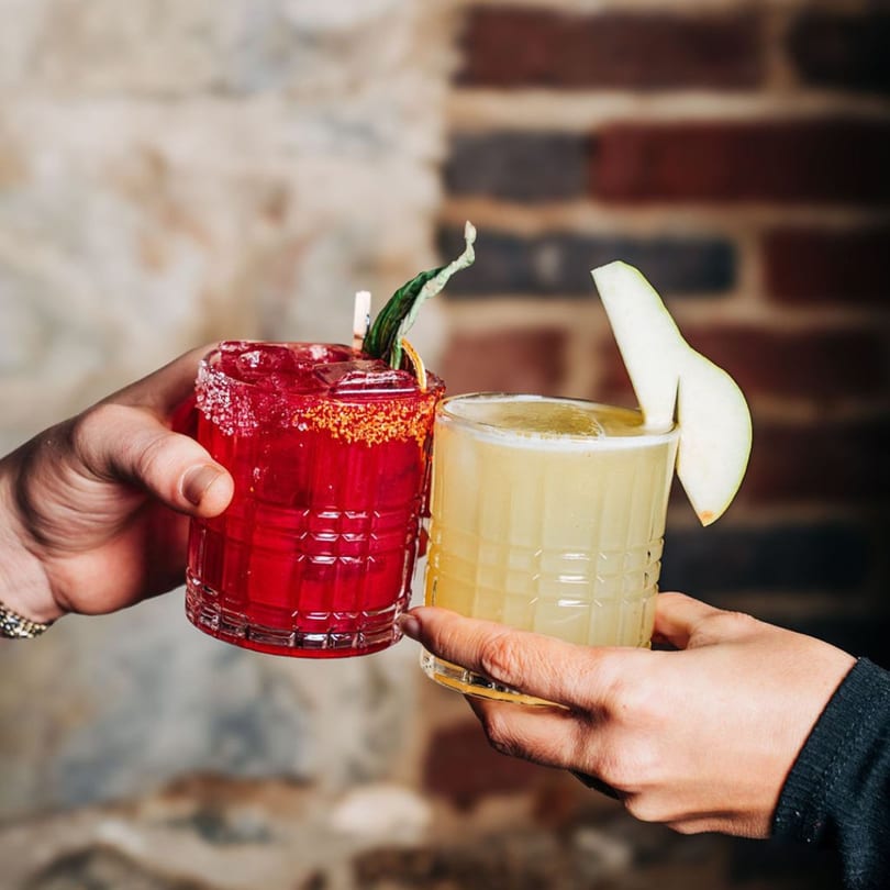Two hands clinking glasses of red and yellow cocktails, garnished with fruit and herbs.