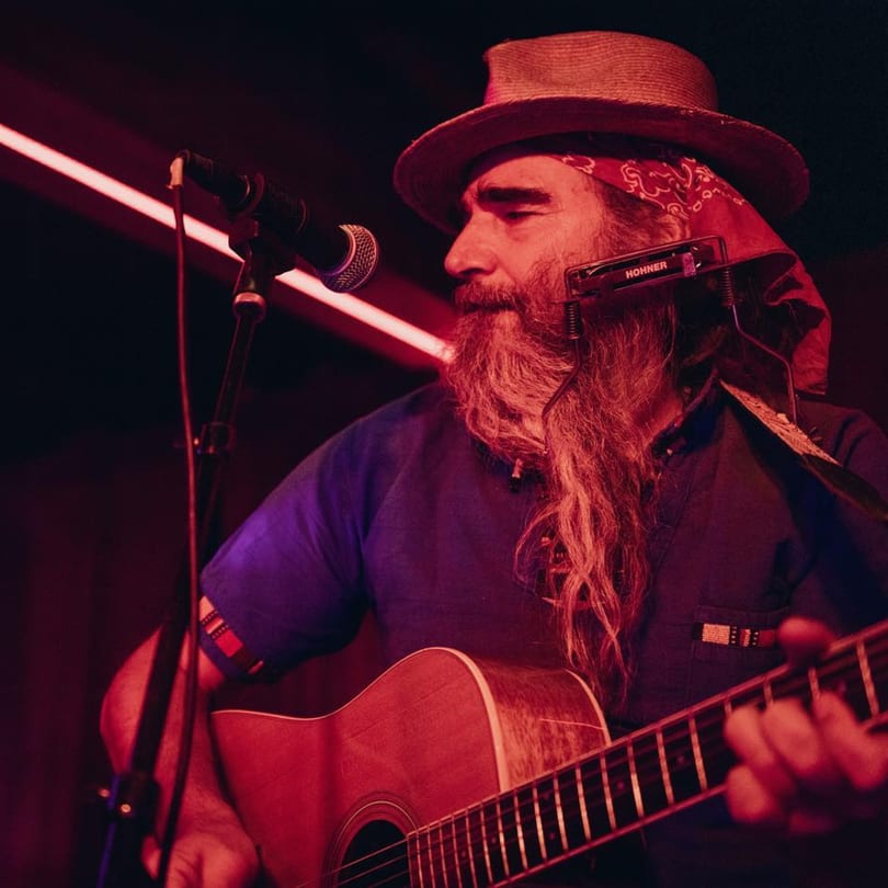 Musician with a long beard, hat, and harmonica playing an acoustic guitar under red lighting.