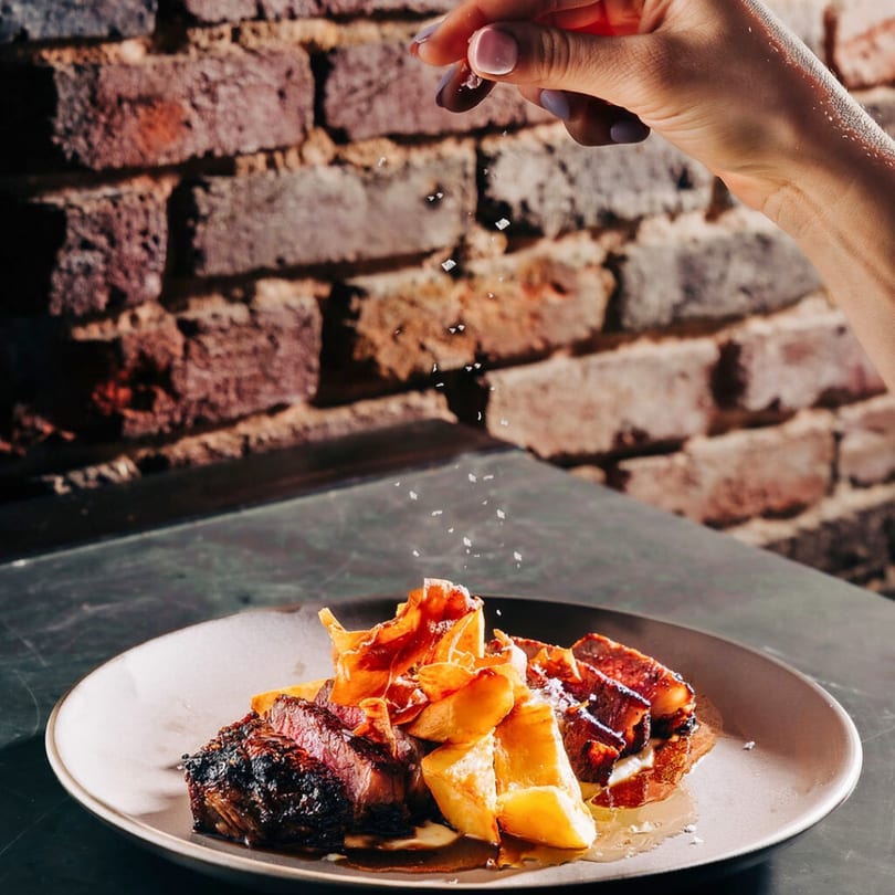 A hand sprinkles salt over a Stone House New American dish of steak, potatoes, and crispy toppings.